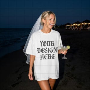 May include: White t-shirt with the text "YOUR DESIGN HERE" in a gothic font. The person is wearing a veil and holding a cocktail with a lime wedge. The background shows a beach at dusk.