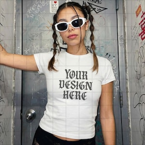 以下が含まれることがあります： ゴシック体の「YOUR DESIGN HERE」の文字が入った白いTシャツ。モデルは白いサングラスをかけ、二つ編みの髪型をしています。背景は落書きで覆われた壁です。