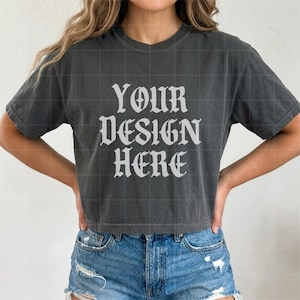 May include: A charcoal grey short-sleeved t-shirt featuring the white text YOUR DESIGN HERE in a gothic-style font. The shirt has a crew neckline and a relaxed fit, paired with blue denim shorts that have a distressed hem and visible stitching.