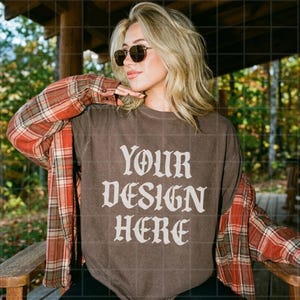 以下が含まれることがあります： ゴシック調のフォントで「YOUR DESIGN HERE」と白文字でプリントされたダークブラウンのクルーネックTシャツ。赤、オレンジ、茶色のチェック柄の長袖フランネルシャツを羽織り、ゴールドフレームのティアドロップ型サングラスを着用している。