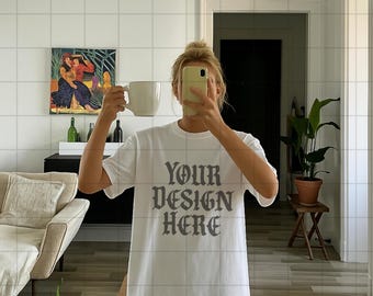 May include: White t-shirt with the text "YOUR DESIGN HERE" in a dark gray, gothic-style font. The person is holding a white mug and a phone. A painting hangs on the wall in the background.