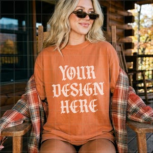 May include: A burnt orange crewneck t-shirt featuring the text YOUR DESIGN HERE in a cream-coloured gothic font. The shirt is worn with a red, white, and green plaid flannel button-down shirt layered over the shoulders.