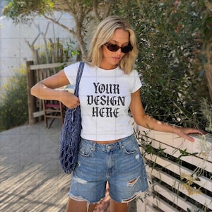 May include: White crop top with the text "YOUR DESIGN HERE" in a gothic font. The model is wearing blue denim shorts and sunglasses. A blue mesh tote bag is over the shoulder.