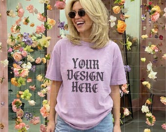 Pode incluir: Uma t-shirt lilás clara com o texto "YOUR DESIGN HERE" em fonte gótica. A pessoa está usando óculos de sol e jeans azuis. O fundo é uma exibição floral com flores rosa e amarelas.