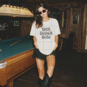 Op de afbeelding: Wit t-shirt met de tekst "YOUR DESIGN HERE" in een gotisch lettertype. De persoon draagt een zwarte korte broek en zwarte cowboylaarzen. De achtergrond omvat een pooltafel en een houten interieur.