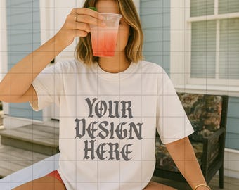 May include: A white t-shirt with the text "YOUR DESIGN HERE" in a dark gray, stylized font. The person is holding a clear plastic cup with a red beverage and a lime wedge. The person is wearing a gold bracelet.