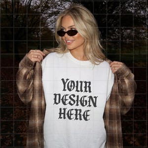 Könnte beinhalten: Ein weißes T-Shirt mit dem schwarzen Text "YOUR DESIGN HERE" in einer gotischen Schriftart, getragen unter einem aufgeknöpften braun-beigen Flanellhemd mit Karomuster. Das Kleidungsstück wird mit einer dunklen, ovalen Sonnenbrille kombiniert.