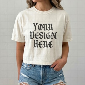 May include: A cream-colored short-sleeve t-shirt featuring the dark gray text "YOUR DESIGN HERE" printed in a gothic-style font on the chest. The shirt has a crew neckline and a straight hem, paired with blue denim shorts with distressed details.