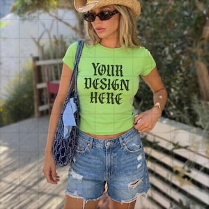 Könnte beinhalten: Ein limettengrünes Crop-Top mit dem Text "YOUR DESIGN HERE" in schwarzen gotischen Buchstaben. Das Oberteil wird mit zerrissenen Jeansshorts kombiniert. Die Person trägt eine Sonnenbrille und einen Strohhut und trägt eine blaue Tragetasche.