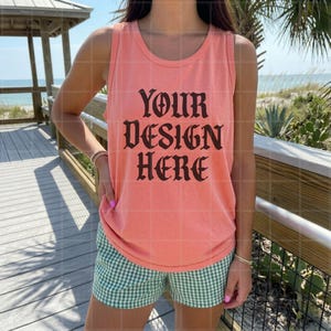 May include: Coral-colored tank top with the text "YOUR DESIGN HERE" in a gothic font. The tank top is paired with green and white checkered shorts. The image is taken outdoors, with a beach and wooden deck in the background.