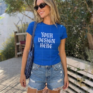 May include: A royal blue t-shirt with the text "YOUR DESIGN HERE" in white, paired with denim shorts. The model is wearing sunglasses and carrying a blue tote bag. The background features greenery and a wooden fence.