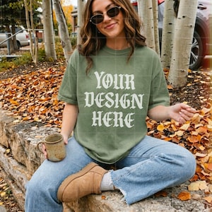 May include: An olive green short-sleeve t-shirt featuring the white text "YOUR DESIGN HERE" in a bold, stylized font. The shirt has a crew neckline and a relaxed fit, worn with light blue denim jeans and tan suede ankle boots.