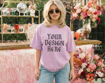Pode incluir: Uma t-shirt lilás clara com o texto "YOUR DESIGN HERE" em fonte gótica. A pessoa está usando jeans azuis e óculos de sol. O fundo inclui um bar com vidraria e arranjos florais.