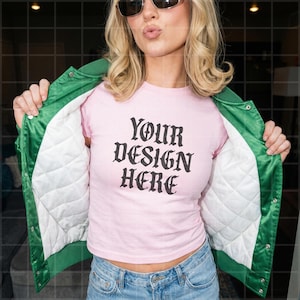 May include: A light pink t-shirt with the text "YOUR DESIGN HERE" in black gothic font. The shirt is worn under a green satin jacket with white quilted lining. The model is wearing blue jeans and sunglasses.