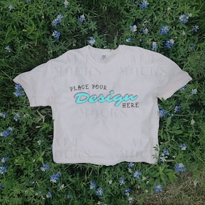May include: A white t-shirt with a distressed look, lying on a bed of blue flowers. The shirt has the text "Place your Design Here" printed on it in a retro font.