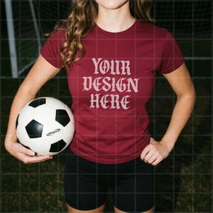 Può includere: Una t-shirt a maniche corte color bordeaux con la scritta "YOUR DESIGN HERE" in un carattere gotico rosa chiaro, abbinata a pantaloncini sportivi neri e a un pallone da calcio bianco e nero con la parola "SPORTS" su un pannello.