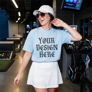 May include: A light blue t-shirt with the text "YOUR DESIGN HERE" in a gothic font. The shirt is paired with a white skirt and a white baseball cap. The person is wearing sunglasses.