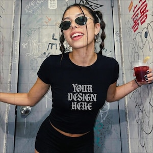 May include: Black crop top with the text "YOUR DESIGN HERE" in a gothic font. The model is wearing black sunglasses and black shorts. The background is a wall with graffiti.