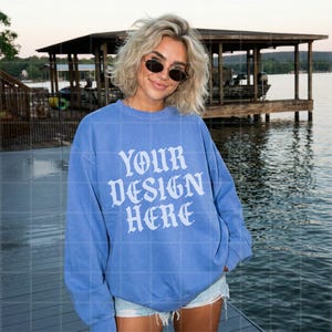 May include: A blue sweatshirt with the text "YOUR DESIGN HERE" in white, displayed on a person. The sweatshirt is a solid blue color. The background includes a lake and a wooden structure.