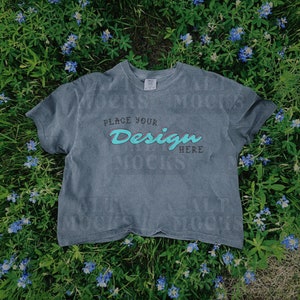 May include: A gray cropped t-shirt with short sleeves lies on a bed of blue flowers and green grass. The shirt has the text "Place Your Design Here" printed on the front.