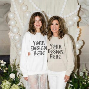 May include: Two people wearing matching sweatshirts with the text "YOUR DESIGN HERE" in a gothic font. One sweatshirt is white, the other is beige. The background includes white balloons and floral arrangements.