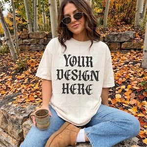 Può includere: Una t-shirt a maniche corte color bianco sporco con il testo nero YOUR DESIGN HERE in un carattere in stile gotico, abbinata a jeans blu, stivaletti in camoscio beige, occhiali da sole scuri e una tazza in ceramica marrone maculata.