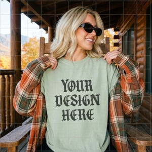 May include: A light green crewneck sweatshirt featuring the black text YOUR DESIGN HERE in a gothic font, layered under an orange, green, and white plaid flannel shirt. The garment is worn with black sunglasses.