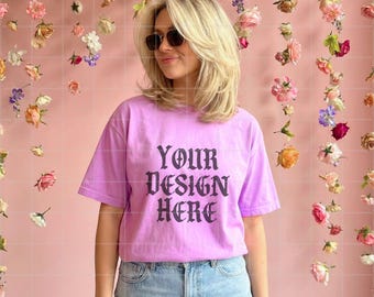 Pode incluir: Uma camiseta lilás com o texto "YOUR DESIGN HERE" em fonte gótica. A modelo usa óculos de sol e jeans azuis. O fundo é uma parede rosa com flores penduradas.