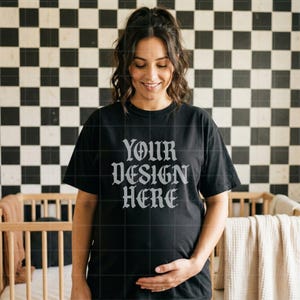May include: Black t-shirt with the text "YOUR DESIGN HERE" in a gothic font. The shirt is worn by a person. The background features a black and white checkered pattern and a wooden crib.