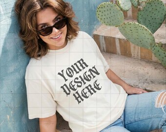 May include: A cream-coloured t-shirt with the text "YOUR DESIGN HERE" in a gothic font. The person is wearing sunglasses and light blue jeans with ripped knees. A cactus is in the background.
