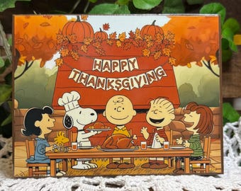 May include: A rectangular sign featuring a Peanuts Thanksgiving scene with Snoopy, Charlie Brown, and friends at a dinner table. The text HAPPY THANKSGIVING is displayed on a banner above a red doghouse, surrounded by orange autumn leaves.