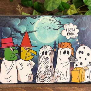 May include: A rectangular wooden sign featuring a Halloween illustration of five figures in costumes against a full moon and dark forest background. The text I GOT A ROCK appears in a speech bubble above a figure wearing a white sheet with black dots.