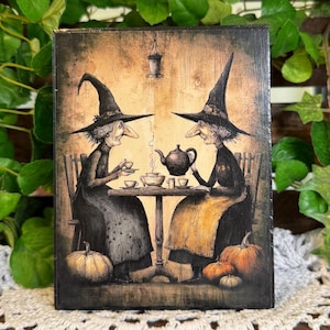 May include: A rectangular wooden wall art piece featuring a detailed illustration of two witches in pointed hats sitting at a table drinking tea. The scene includes a teapot, cups, and pumpkins in a warm, sepia-toned, sketch-like artistic style.
