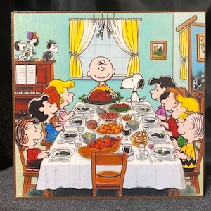 May include: A wooden block featuring a Thanksgiving scene with Charlie Brown, Snoopy, and other Peanuts characters gathered around a table laden with food. The artwork is colorful, with a focus on the characters and the festive meal.