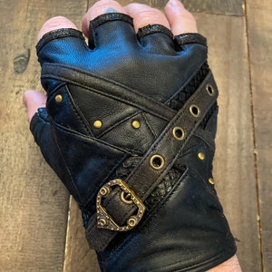 May include: Black leather fingerless gloves with brown straps and metal accents. The gloves have a criss-cross design and a buckle closure.