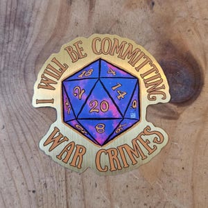 May include: A gold-coloured sticker featuring the text "I WILL BE COMMITTING WAR CRIMES" around a blue and pink twenty-sided die. The die has white numbers and is centrally positioned.