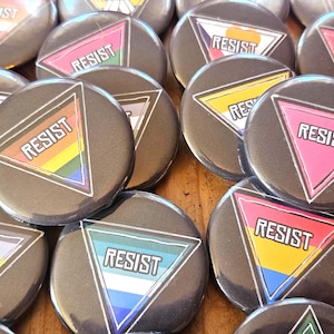 May include: A collection of round, dark gray buttons with the word "RESIST" inside a triangle. The triangles feature various rainbow and pride flag color schemes. The buttons are arranged on a wooden surface.