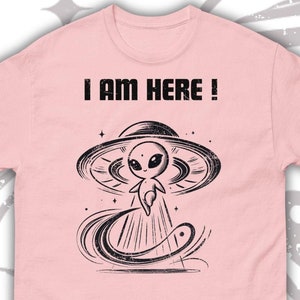 以下が含まれることがあります： ピンクのTシャツに、UFOに乗った宇宙人の白黒イラストが描かれています。Tシャツには「I AM HERE！」の文字がプリントされています。