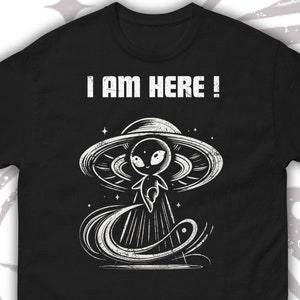 以下が含まれることがあります： UFOに乗った宇宙人の白いグラフィックが描かれた黒いTシャツ。Tシャツには「I AM HERE！」という文字が印刷されています。