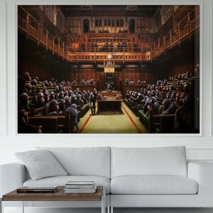 Op de afbeelding: Een ingelijst kunstwerk dat een parlementszaal vol chimpansees afbeeldt. Het schilderij heeft gedetailleerde architectonische elementen en een levendig kleurenpalet, met de primaten zittend in de parlementaire banken. Het kunstwerk wordt tentoongesteld boven een witte bank en een houten salontafel.
