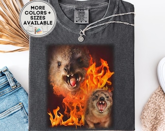 May include: A charcoal gray t-shirt featuring a graphic print of two quokkas with open mouths surrounded by bright orange and yellow flames. The shirt has a crew neckline and a Comfort Colors label at the collar. A circular badge reads MORE COLORS + SIZES AVAILABLE.
