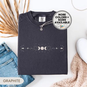 May include: Graphite-colored sweatshirt with a crew neck and a celestial design featuring moon phases and arrows. The shirt has a relaxed fit and is made of a soft material. The text "MORE COLORS + SIZES AVAILABLE" is in a circle on the top right.