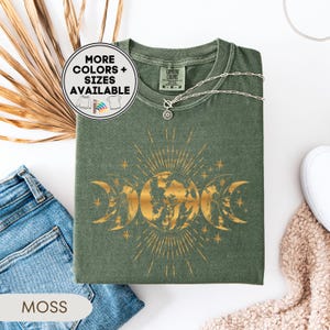 May include: Moss green t-shirt with a gold graphic of the earth, moons, and sunburst design. The shirt has a crew neck and short sleeves. A silver necklace with a sun pendant is on the shirt. The words "MORE COLORS + SIZES AVAILABLE" are in a circle on the shirt.