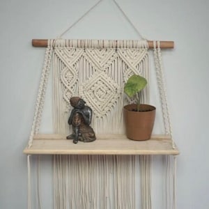 Puede incluir: Un tapiz de macramé de pared con un estante de madera. El estante está decorado con una pequeña estatua y una planta en maceta.