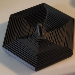 May include: A black hexagonal object with a layered design and a small, black, cylindrical knob on top.
