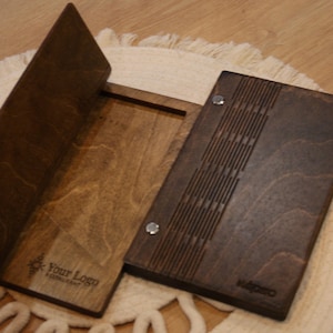 May include: Wooden restaurant menu holder with a dark brown finish. The menu holder is open, revealing the interior. The cover features a decorative design and the text "Your Logo Restaurant".
