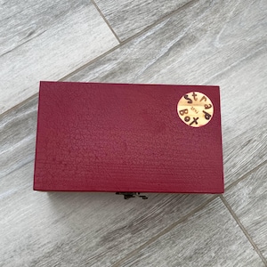 May include: A rectangular, claret-coloured wooden box with a textured surface. A small, round, wooden sticker with the text "strap box" is affixed to the top. The box is closed with a metal clasp.
