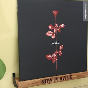 Może przedstawiać: Czarna płyta winylowa z czerwonym wzorem róży i tekstem "violator" na okładce. Płyta znajduje się na drewnianej półce z wygrawerowanym tekstem "NOW PLAYING". Płyta jest Depeche Mode.