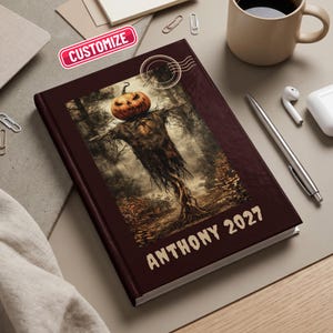 May include: A dark maroon hardcover journal featuring a central rectangular illustration of a scarecrow with a carved pumpkin head in a forest. The text ANTHONY 2027 is printed in a light tan font at the bottom, with a circular postmark graphic above.
