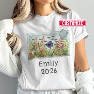 Puede incluir: Camiseta blanca con una ilustración de acuarela de un acuario con peces, plantas y un castillo. El texto "Emily 2026" está debajo. Una pancarta roja "Personalizar" está en la parte superior.